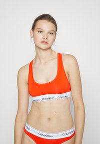 Fényes narancssárga sportmelltartó széles pántokkal, amelyen egy fehér elasztikus szalag található "Calvin Klein" márkázással. Simaszövetű és hozzáillő fazonú.
