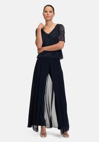 Granatowa koronkowa bluzka z krótkimi rękawami, dekoltem w serek, nałożona na przewiewny kombinezon z szyfonu z rozcięciem z przodu i przezroczystą wstawką, w połączeniu z srebrnymi szpilkami.