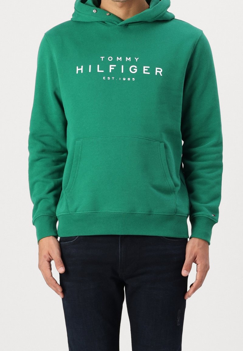 Homme portant un sweat à capuche vert Tommy Hilfiger avec poche avant et texte du logo en blanc, associé à un jean bleu foncé.