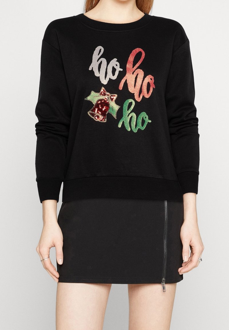 Femme portant un sweat-shirt noir avec un texte scintillant "ho ho ho" et un motif de cloche rouge, associé à une jupe noire avec une fermeture éclair à l'avant.