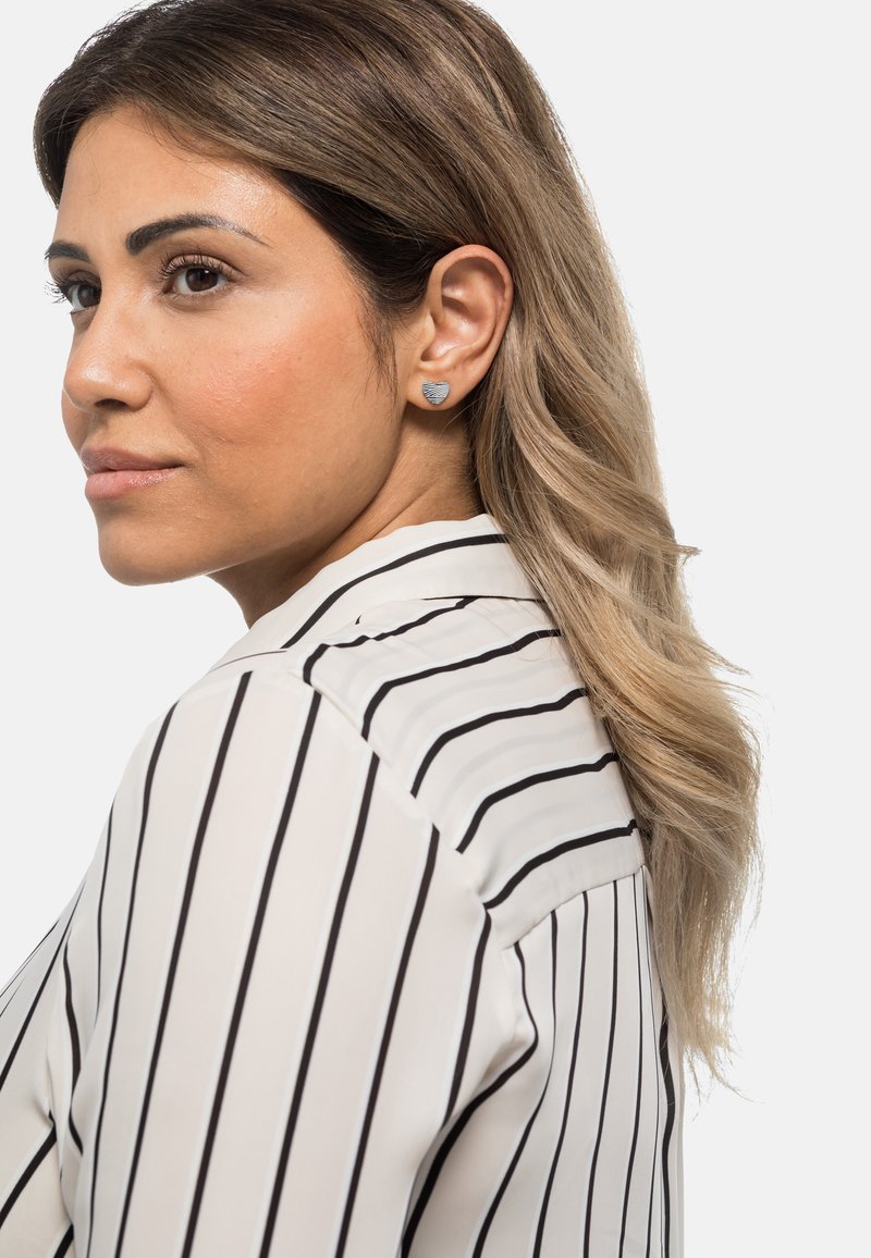 Boucles d'oreilles en argent en forme de cœur portées par une personne, sur fond blanc. La chemise est à rayures verticales noires et blanches.