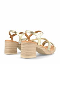 Sandalias de tacón de metal dorado con tiras entrelazadas, un tacón de bloque de madera y una suela de goma texturizada. Presenta un suave brillo.