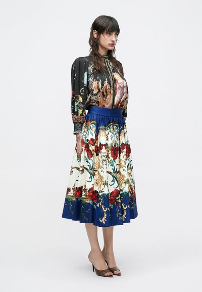 Naine kannab pikkade lilleliste seelikut sinise äärega ja musta pluusi, millel on renessansstiilis maalitud figuurid, jalas kõrge kontsaga sandaalid.