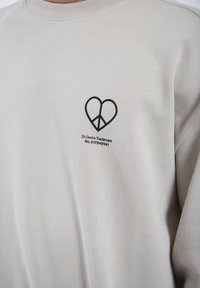Felpa grigia chiara con maniche lunghe e collo rotondo, caratterizzata da un logo a forma di cuore nero e simbolo della pace sulla parte sinistra del petto.