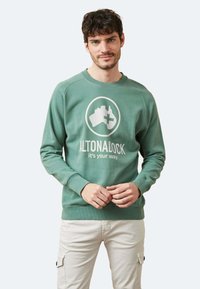 Sudadera verde hecha de una mezcla de algodón, con un logo redondo que muestra la forma de Australia y texto, puños acanalados y dobladillo. Llevada sobre pantalones claros.