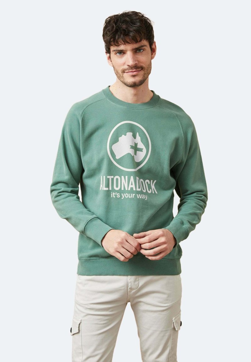 Sudadera verde hecha de una mezcla de algodón, con un logo redondo que muestra la forma de Australia y texto, puños acanalados y dobladillo. Llevada sobre pantalones claros.