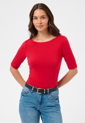 Femme aux longs cheveux bruns porte un haut rouge ajusté à manches mi-longues, rentré dans un jean bleu avec une ceinture noire sur un fond blanc uni.