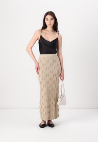 Schwarzes Satin-Camisole mit dünnen Trägern, kombiniert mit einem beigen, strukturierten Maxirock mit Fransenmuster, ergänzt durch eine weiße Handtasche und schwarze Ballerinas.