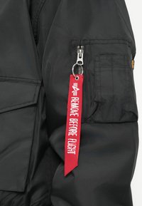Giacca nera in nylon con una tasca laterale con zip e un'etichetta rossa di tessuto che recita "RIMUOVERE PRIMA DEL VOLO". Presenta una superficie testurizzata e dettagli di cucitura.