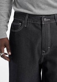 Jeans en denim noir avec des coutures blanches, comportant une poche avant et une fermeture à bouton. Le tissu a une texture lisse.