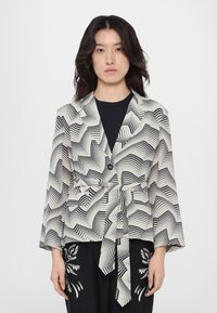 Blazer a righe in bianco e nero realizzato in un tessuto leggero, con vita legata e maniche ampie, caratterizzato da un design a zigzag e chiusura con un singolo bottone.