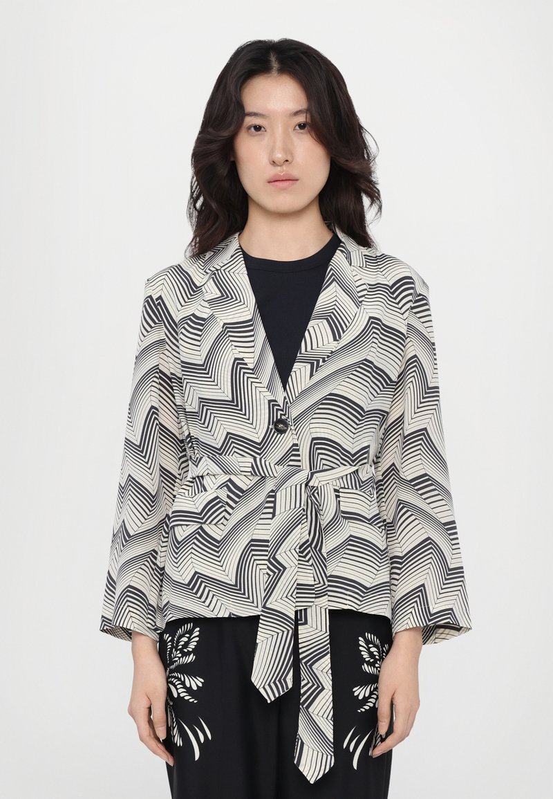 Blazer a righe in bianco e nero realizzato in un tessuto leggero, con vita legata e maniche ampie, caratterizzato da un design a zigzag e chiusura con un singolo bottone.