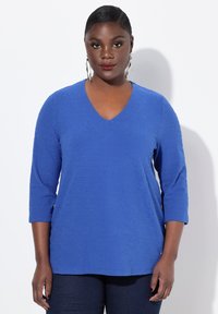 Top azul de cuello en V con un patrón geométrico texturizado, mangas tres cuartos y un corte relajado. Combinado con pantalones de mezclilla oscuros.
