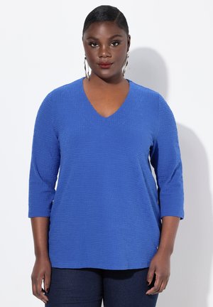 Blauwe V-hals top met een textuur van een geometrisch patroon, driekwartmouwen en een relaxte pasvorm. Gecombineerd met donkerdenim broek.