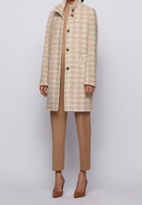 Kvinde iført beige hundetandsmønstret frakke med knapper over kamelfarvede bukser og matchende højhælede sko, stående mod en ensfarvet baggrund.