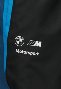 Μαύρο υφασμάτινο πάνελ με λευκό στρογγυλό λογότυπο BMW, τριπλό ριγωτό λογότυπο "M" και κείμενο "Motorsport", δίπλα σε μπλε υφασμάτινη επιφάνεια.