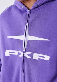 Sudadera púrpura con cremallera en la parte delantera, que presenta un gráfico de estrella blanca y la letra "PXP". Material suave con una textura lisa.