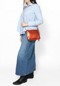 Sac bandoulière orange métallique au design structuré, doté d'une chaîne et d'accents angulaires, tenu par une personne en tenue décontractée.