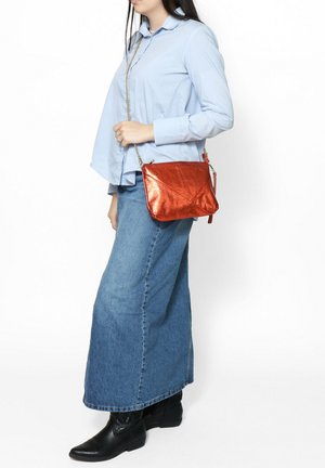 Borsa a tracolla metallica arancione con un design strutturato, dotata di una tracolla a catena e dettagli angolari, tenuta da una persona in abbigliamento casual.