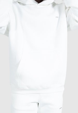 Osoba ubrana w prostą białą bluzę z kapturem i dopasowane białe spodnie dresowe, z podniesionymi rękami, bez widocznych szczegółów tła.