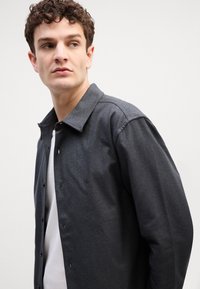 Camicia button-up grigio scuro con colletto a punta, realizzata in un tessuto testurizzato e con un sottile dettaglio del logo, indossata sopra una t-shirt bianca.