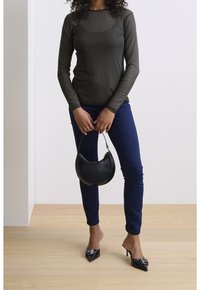 Femme portant un haut noir transparent à manches longues, un jean skinny bleu, des escarpins noirs à bouts pointus, tenant un petit sac à main noir par la poignée.