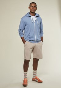 Hellblaue Zip-Hoodie mit weißen Kordeln, beigen Shorts, gestreiften Socken und orangenen Sneakers mit türkisfarbenen Akzenten. Einfaches Design, lässige Passform.