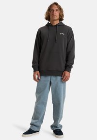 Dark gray hoodie with a front pocket, adjustable drawstrings, and logo on the chest. Paired with light blue, loose-fitting jeans and navy shoes.
