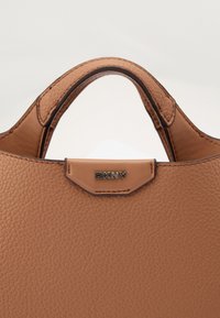 Világosbarna bőrtáska texturált felülettel, lekerekített felső résszel, két fogantyúval és egy kis fém DKNY logóval az elején.