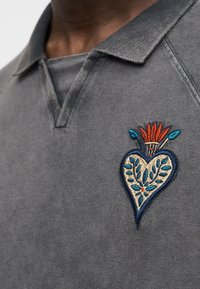 Κοντινό πλάνο γκρι πόλο πουκάμισου με πολύχρωμο κεντητό σχέδιο καρδιάς και λουλουδιών στην αριστερή πλευρά του στήθους.