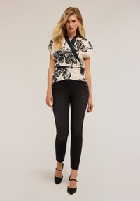 Mujer con cabello rubio que lleva una blusa envolvente color crema con estampado floral negro, pantalones skinny negros de tiro alto y tacones negros puntiagudos.