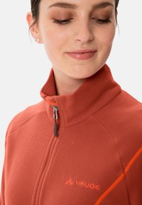 Punane fliistekas, millel on kõrge krae ja oranžid aktsendid. Omab lukku tõmblukuga ja VAUDE logo rinnal. Pehme tekstuur.