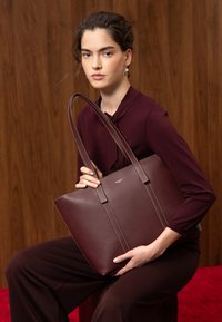Femme portant une tenue bordeaux, assise et tenant un grand sac cabas en cuir bordeaux assorti devant un fond en bois.