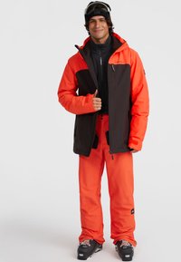 Heldere oranje geïsoleerde jas met zwarte accenten, ritszakken en een hoge kraag. Bijpassende oranje skibroek voltooit de outfit, met skischoenen.