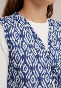 Sinine vatitatud vest valge geomeetrilise mustriga, millel on sidumisakt, ümar krae ja tekstuuriga kangas. Kantud valge särgi peal.