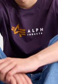 Fialové bavlněné tričko s výrazným žlutým a bílým logem "ALPHA" s křídlatým designem. Materiál má hladkou texturu.