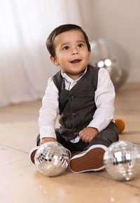 Grijze visgraatvest over een wit shirt, zittend op een lichte vloer. Houdt twee zilveren discoballen vast, met bruin-gevoerde witte schoenen zichtbaar.