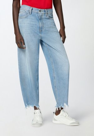 Osoba ubrana w jasnoniebieskie jeansy z postrzępionymi nogawkami, białe trampki i czerwony top, stojąca na tle jednolitego białego tła.