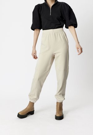 Femme portant un haut noir à manches bouffantes avec fermeture éclair, un pantalon de jogging crème à taille élastique, et des bottines à plateforme beige sur fond blanc.
