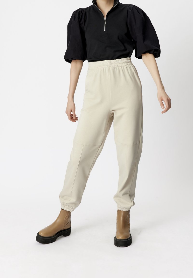 Femme portant un haut noir à manches bouffantes avec fermeture éclair, un pantalon de jogging crème à taille élastique, et des bottines à plateforme beige sur fond blanc.