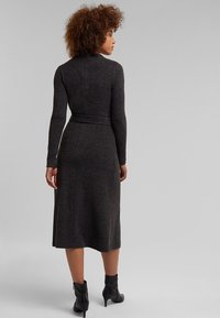 Femme aux cheveux bouclés portant une robe midi à manches longues de couleur gris foncé et des bottines noires, debout de dos face à la spectatrice.