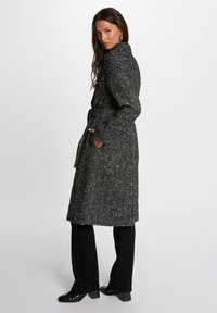 Zwart-wit gemêleerde jas met een riem, hoge kraag en zijzakken. Gecombineerd met zwarte flared broek en glanzende zwarte schoenen.