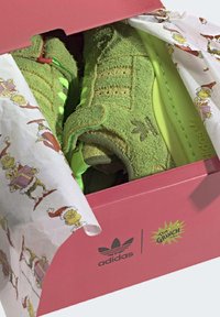 Zelené texturované tenisky Adidas uvnitř růžové krabice s vánočním papírem a logem na téma Grinch.