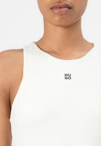 Hvit ermeløs topp med rund hals; har en liten svart "HUGO" logo på brystområdet. Glatt stoff og tettsittende design.