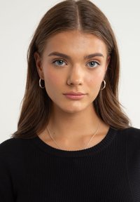 Schwarzer gerippter Pullover mit Rundhalsausschnitt, getragen mit einer silbernen Halskette. Das Model hat mittelbraunes Haar und trägt Creolen.
