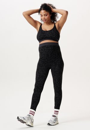 Zwangere vrouw draagt een zwarte sport-bh met luipaardprint en legging, witte sokken en grijze sneakers, poseert met handen op het hoofd.