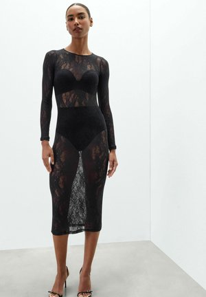 Femme portant une robe midi en dentelle noire à manches longues avec des sous-vêtements noirs visibles, debout contre un mur blanc uni.