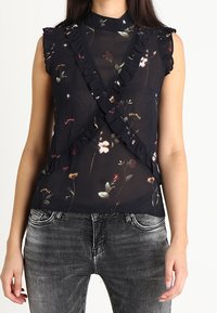 Black sheer top with floral patterns, ruffled accents along the shoulders and front, high neckline, paired with grey jeans.