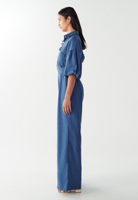 Combinaison en denim bleue, avec un col à revers, des manches courtes bouffantes et un design à jambes larges. Accentuée de boutons sur le devant et d'une taille ceinturée.