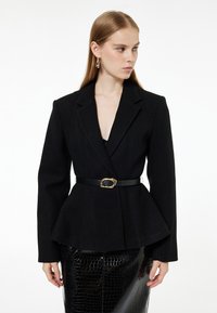 Czarna blazerka peplum z teksturowanej tkaniny, z dopasowaną talią i czarnym paskiem z złotymi akcentami. Połączona z błyszcząca, wzorzystą spódnicą.